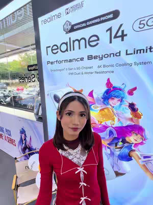 A woman in a red top stands in front of a large poster promoting a Realme 14 Series phone. The poster displays the phone's name in bold, along with graphics and some of its features. The woman is smiling and the background shows signs of a retail setting.