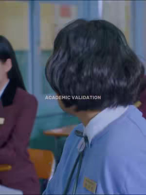 The image shows a person facing away from the camera. The person is wearing a blue sweater over a white collared shirt. The background appears to be inside a classroom with another person partially visible on the left wearing a red jacket. The added text overlay says "ACADEMIC VALIDATION".