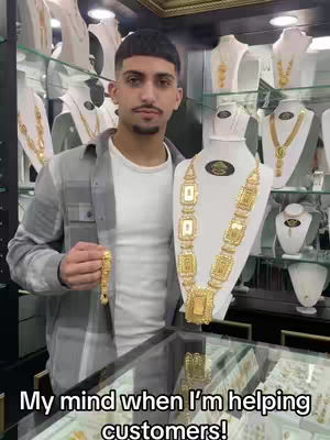 A man stands in a jewelry store holding a bracelet, with a large gold necklace displayed nearby on a bust. Various necklaces and jewelry are visible in the background. Below on a text overlay, the phrase 'My mind when I'm helping customers!'