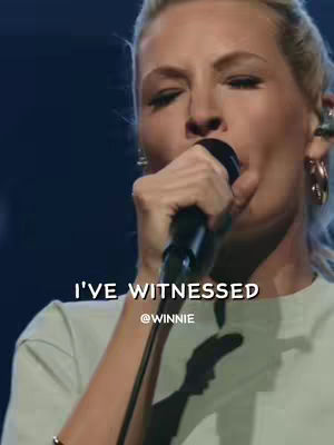 A woman with blonde hair is singing into a microphone. The background appears to be a stage due to the dark blue lighting. Text overlays on the image. The woman wears a light colored shirt and hoop earrings.