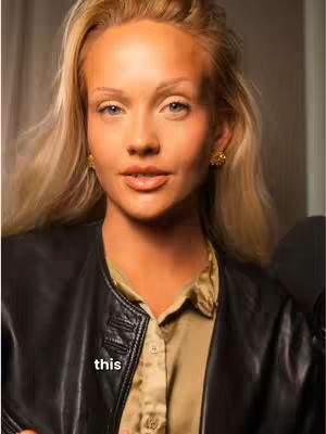 A close-up shot of a fair-skinned woman with long, blonde hair. She's wearing a black leather jacket over a gold-colored blouse. The woman has a slight smile, light eye color, and is wearing gold earrings. The image has a warm color tone, with soft lighting and subtle shadows.