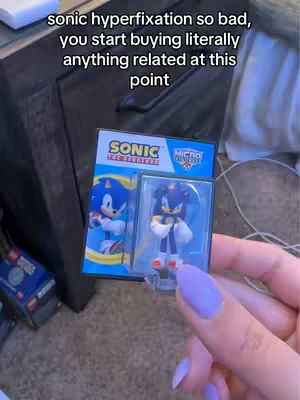 A hand holding a box containing a Sonic the Hedgehog figure. The background shows a dark wooden surface, a rug, and a white sheet. The hand has purple nail polish. Text overlayed at the top includes “sonic hyperfixation so bad, you start buying literally anything related at this point”.