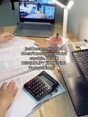A well-lit workspace features a laptop displaying a video, a calculator, papers with handwriting, a pink mug with a spoon, and a desk lamp. Text overlays provide motivation and encouragement.