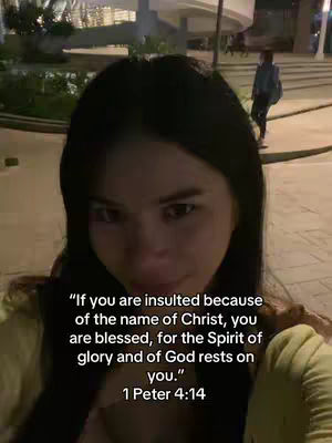 A selfie-style video cover image features a woman with dark hair and a somewhat serious expression. She is positioned from the chest up, with the background slightly blurred, showing outdoor elements. The lighting appears to be soft and natural. Overlayed on the image is a Bible verse, written in white against the background.