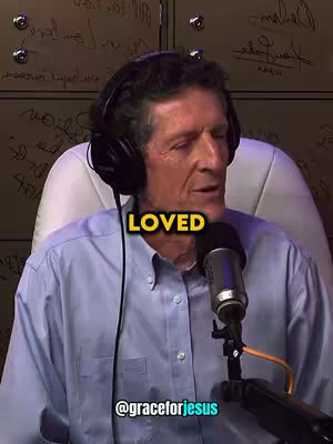 A middle-aged man with short brown hair, a light blue button-down shirt, and a black headset is the central focus, positioned against a backdrop covered in what appears to be handwritten notes and signatures. The word "LOVED" is displayed in a yellow box above the man's head, and the bottom of the image includes a watermark with the username "@graceforjesus". The lighting is moderate and suggests an indoor setting, likely a studio, with a microphone positioned in front of the man.
