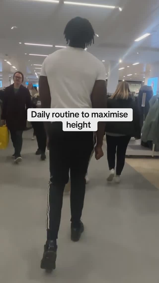 A man walks towards the camera in a shopping center. He is wearing all black athletic pants with a white stripe on the side, a white t-shirt and black shoes. The background shows multiple people and store shelves.