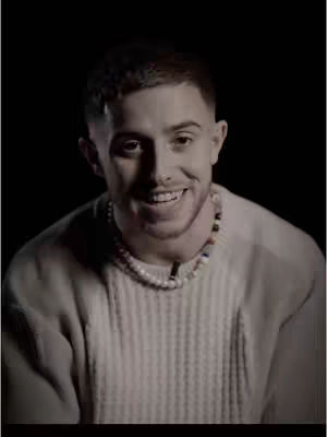 A man with short brown hair is centered in front of a dark backdrop. He has a light complexion and is smiling widely, showing his teeth. He wears a cream-colored knit sweater and a beaded necklace. The lighting is focused on his face, creating a contrast with the shadowed background.