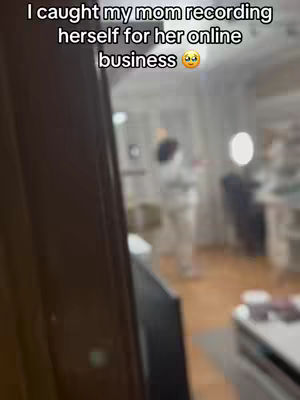 The image is blurred, showing a room interior with a partially visible door frame on the left. A woman is standing further into the room, out of focus. The text overlay at the top reads: I caught my mom recording herself for her online business.