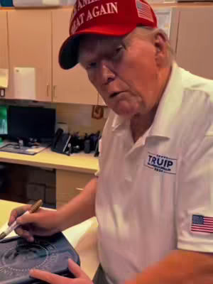 The image shows Donald Trump in a room, wearing a red 'Make America Great Again' hat and a white polo shirt with 'TRUIP 2024' printed on it, and the American flag on the sleeve. He is holding a pen and appearing to sign a book. The background consists of a wood-paneled wall and shelves.