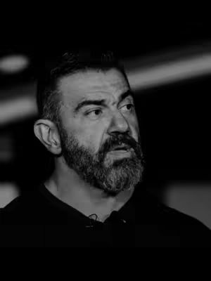 A black and white image of a man with a beard looking off to the side. The man is wearing a dark shirt, and the background is shadowy.