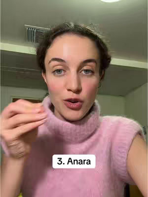 A woman with fair skin and brown hair is the central figure, framed in a medium shot. She has blue eyes and slightly parted lips, appearing to be speaking. The immediate foreground is dominated by her pink, high-collared sweater. Behind her, the background is blurred but includes hints of a room with a ceiling and possibly a light fixture or ventilation vent. Below is a text box with the text "3. Anara".