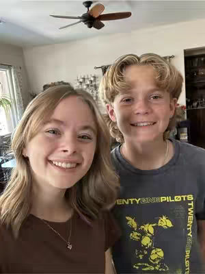 Two young people, a female and a male, are in a selfie style shot in a bright indoor setting. The female is on the left and the male is on the right. They are both smiling at the camera.