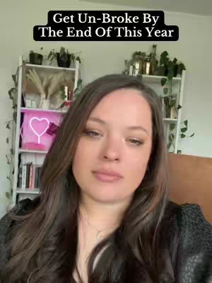 A woman with long brown hair is centered in the frame, looking at the camera with a neutral expression. Above her head is a black banner with white text. Behind her, a white shelf is filled with plants and decorative items.