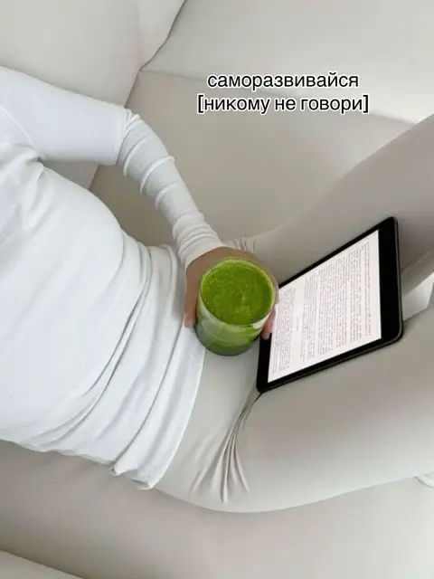 A person in white clothing is lying on a white couch, holding a green smoothie in one hand and a tablet displaying text in the other. The background is plain white; the overall image is minimalist.