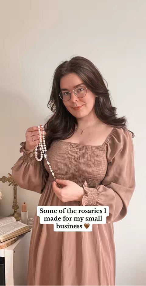 A young woman with long brown hair, glasses, and a cross necklace is centered. She wears a dusty rose dress and holds a rosary. Text overlayed reads: Some of the rosaries I made for my small business.