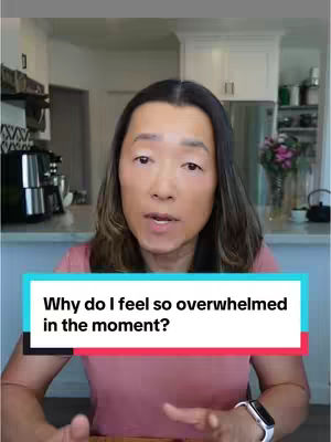 A woman with medium-length dark hair is centered in the frame, looking directly at the camera. She's wearing a pink top. Below her, a text box with a white outline and a blue backdrop highlights the question 'Why do I feel so overwhelmed in the moment?' The background offers a glimpse of a kitchen setting, adding depth to the scene.