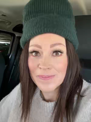 A woman wearing a dark green knitted beanie and a white sweater is sitting inside a car, looking directly at the camera with a slight smile. The background suggests a parking lot or street setting, with the interior of the car partially visible.