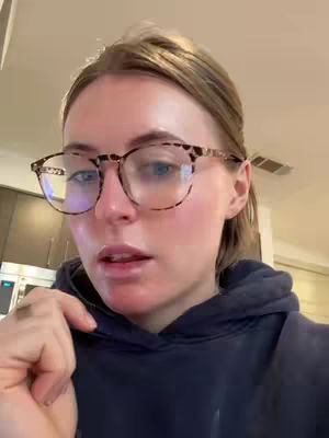 A close-up selfie features a young woman wearing eyeglasses and a dark blue hoodie. The background is blurred, suggesting an indoor setting. The woman gazes towards the lower left, offering a direct yet pensive look. The lighting appears natural, highlighting her features. 