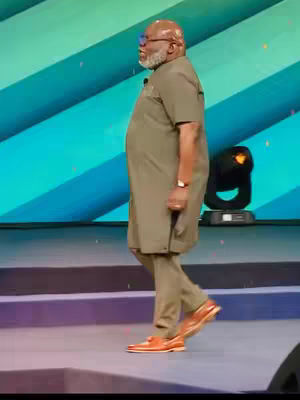 The image shows a man in a medium shot, walking across a stage. He has a bald head and a gray beard, suited in a long-sleeved, olive-green suit and matching pants. He has tan leather shoes, walking towards the left side of the frame on a purple platform, with a light spotlight in the background. The backdrop is a teal and blue gradient color scheme.
