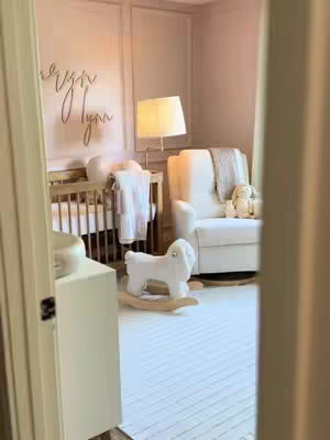 An interior shot framed through a doorway shows a baby's nursery. The space is bathed in warm, neutral tones, with a crib, rocking chair, and soft lighting creating a cozy atmosphere. The room includes a rug and furniture. The overall impression is one of gentle elegance.