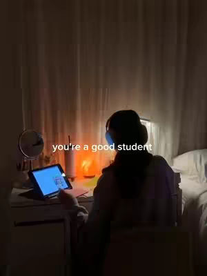 A dimly lit room with a desk where a person sits, lit by a desk lamp and a glowing salt lamp. The person, silhouetted, faces away, with headphones on. On the desk is a tablet with a lit screen; a mirror and pens are also visible. The text overlay states, "you're a good student."