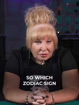 The image features a woman with blonde hair styled in a high bun, wearing a black shirt. Text overlayed on the image reads, "SO WHICH ZODIAC SIGN". The woman appears to be seated with her hands clasped in front of her.