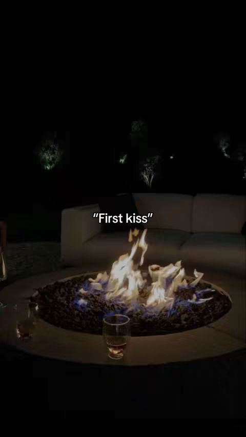 A dimly lit image of a fire pit with flames, surrounded by a dark setting. In the foreground, there are two glasses. Above the flames, the text "First kiss" is displayed. The background shows a white couch and some trees. The overall mood is intimate.