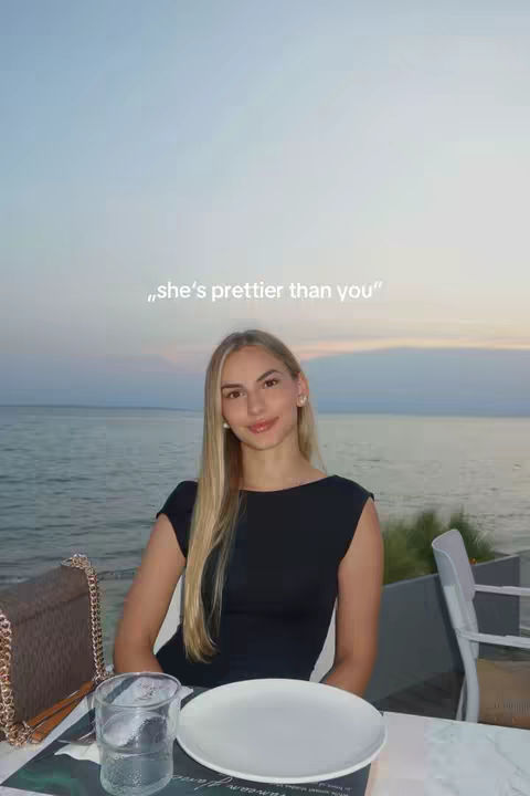 A young woman with long blond hair sits at a table, smiling at the camera. She wears a black dress and is seated outdoors near a body of water. Text overlayed on the image reads, "she's prettier than you."