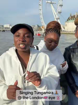 A group of three women are standing by a body of water, with the London Eye and other buildings visible in the background. The woman in the foreground is wearing a white hoodie and a black cap, with a centered text overlay at the bottom of the image. The image is taken during daylight, with a slightly greyish sky.