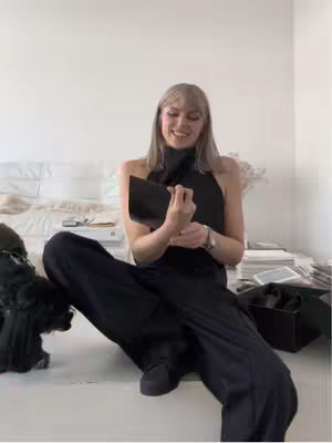 A woman in a black outfit is sitting on a white surface. She is holding a black object. The background is a plain white wall and a bed visible in the background. The composition of the image is well-lit, with an emphasis on the woman's expression.
