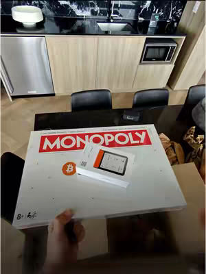 A person unboxes a Ledger Flex Monopoly Bitcoin Bundle. The cover image shows the Monopoly game box lying open on a table, with the Monopoly logo in red text atop a white background. A Ledger Flex device and other materials also lie on top of the box. The scene takes place indoors.