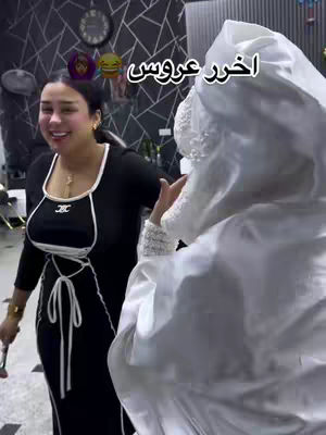 A candid shot showing a woman on the left smiling and partially facing the camera, dressed in a black dress with white trim and gold necklace.  On the right, another person is partially visible, wearing a white bridal veil. The background shows a room with a dark patterned wall, further back is a wall clock.