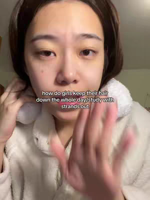 A close-up shot of a young woman's face, with her hands slightly blurred in the foreground. She has dark hair and is wearing a light-colored, fuzzy jacket. The background is a blurry indoor setting. Text overlay questions how girls keep their hair down during study.