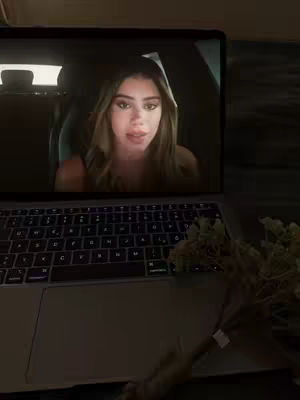 A laptop displays a video of a woman speaking in a car. A bouquet of flowers lies adjacent.