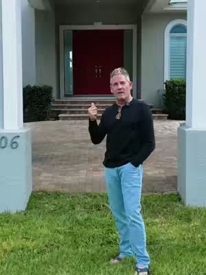 A man stands in front of a large house with a red door, giving a thumbs-up. He wears a black shirt, blue jeans, and sandals. The scene is bright, with green grass and a clear sky suggesting a sunny day.