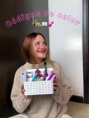 A young woman with blonde and light brown hair holds a white basket filled with colorful pens. The background is split between a dark grey wall on the left and a white wall on the right. Pink text overlayed with a neon effect appears above the woman's head, along with icons and hearts.