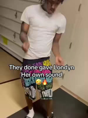 A person in a white t-shirt and colorful shorts is dancing enthusiastically. The image is focused, with the individual centered, and the background showing a hint of a hallway.