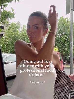 A woman with light skin is posing in front of an outdoor background with a lot of greenery. She's wearing a white sleeveless top and has her arm raised, with her hand near her head. Text overlays the image, and there's a hint of a restaurant setting visible.