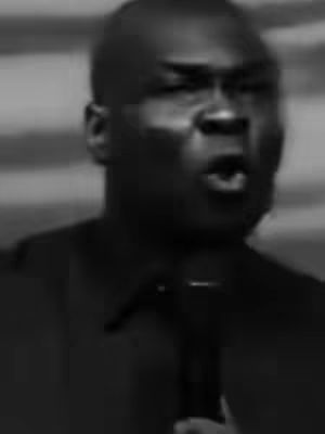 A black and white, medium shot of a bald Black man speaking into a microphone. His facial expression is animated and his mouth is open. The background is blurred. The man is wearing a dark-colored collared shirt.