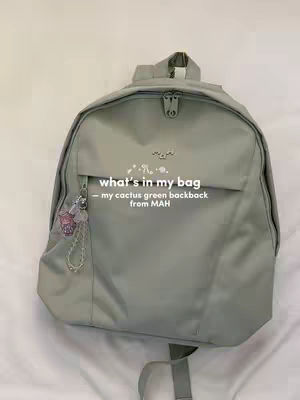 A close-up shot of a sage green backpack sits against a plain off-white background. The backpack is centered in the frame. The top has a handle and a zipper closure. The main body is a single pocket with a smaller pocket on the front side. The front pocket has a charm attached to its zipper. Above this, the text is written in white font. The image is straightforward, focusing on the backpack and its details.