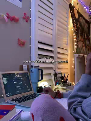A cozy indoor study scene, the composition is focused on the workspace. A laptop sits open, displaying a screen filled with applications, positioned on a desk. A book is visible, alongside a blue insulated water bottle, a pen holder filled with pens, a desk lamp, and some other decorative supplies. The background features white louvered shutters and string lights, and some pink floral decoration.