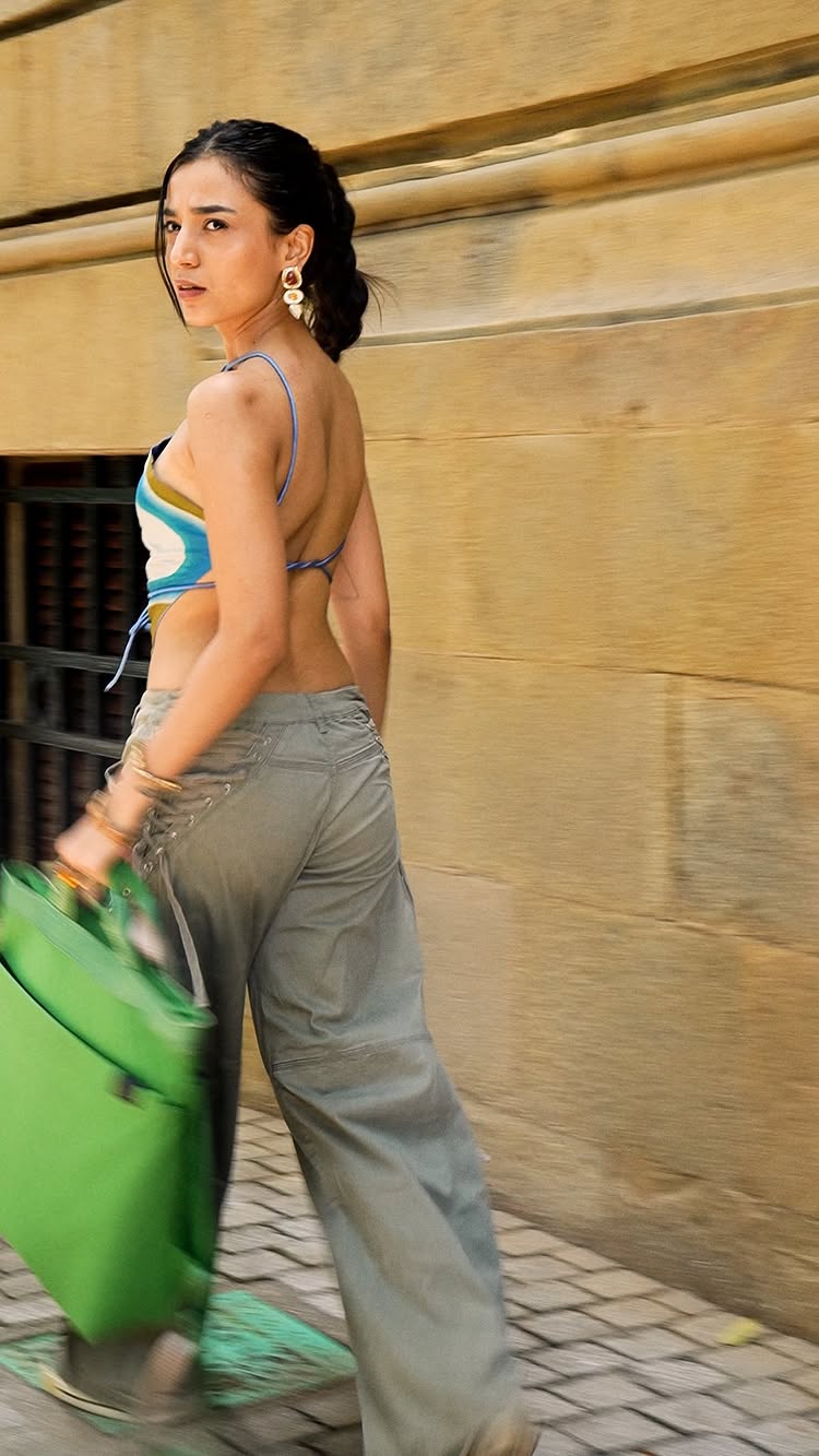 A woman with dark braided hair, large earrings, and dressed in a crop top and cargo pants, walks away from the camera, holding a green bag. The background is blurry with beige-colored walls, and the image has a sense of movement.