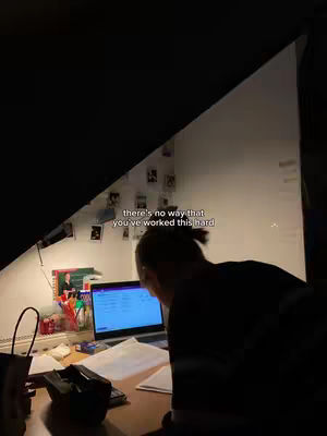 A dimly lit room with a person sitting at a desk, back to the camera, working on a laptop. The wall has some items tacked to it.