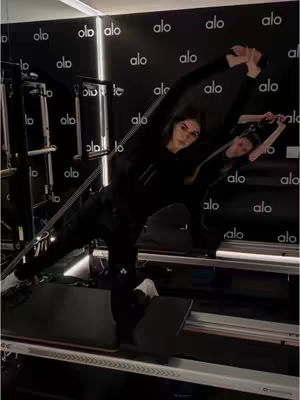 A woman in athletic wear performs a pilates exercise on a machine in a gym. The background features a wall with a repeating logo pattern. The composition is dynamic, with the woman's body at an angle, conveying movement and fitness.