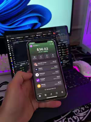 A hand holding a phone displaying a crypto trading app in front of a laptop and a blue-toned computer monitor, with a white keyboard in the background.