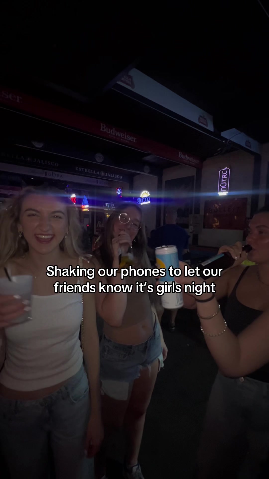 Three young women are shown inside a venue, likely a bar, with bright lights and beer signs visible. They are taking selfies. The woman on the left is smiling, while the woman in the middle is holding a nearly empty can of, NÜTRL. The woman on the right is smiling and holding a microphone.