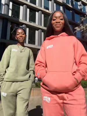 Two young women, likely twins are posed in front of a modern building, dressed in matching, casual outfits. The woman on the left wears a green hoodie and matching pants, while the woman on the right wears a coral hoodie and pants. The background shows a large building with repetitive architectural designs.