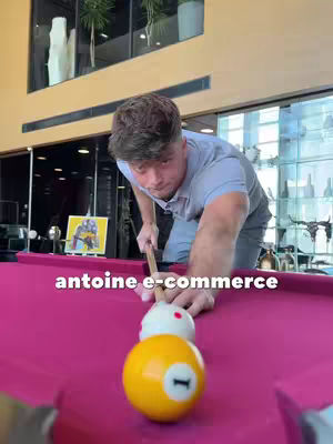 A man is positioned over a pink pool table preparing to hit a billiard ball with a pool cue. The table has two balls in front of him, one white with a red dot and one yellow with the number 1. In the background, there's a vibrant setting.