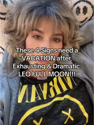 A selfie-style image with a woman at the center, framed from the chest up. She is wearing a black t-shirt with a yellow smiley face design and a gray hooded jacket. The background is blurred, but appears to be a light-colored surface. Text overlayed on the image reads: "These 4 Signs need a VACATION after Exhausting & Dramatic LEO FULL MOON!!!"