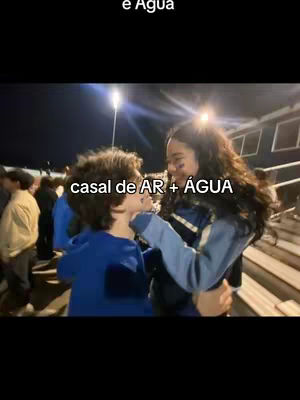 The image shows a couple facing each other, with the woman on the right touching the man's face. They are surrounded by other people and what appears to be bleachers in the background. The text overlay reads 'casal de AR + ÁGUA'.